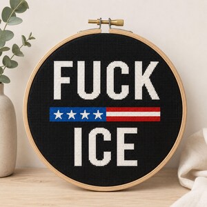 Op de afbeelding: Een ronde houten borduurring met zwarte stof waarop de tekst FUCK ICE in witte kruissteek is geborduurd. Tussen de twee woorden bevindt zich een horizontale balk met vier witte sterren op een blauwe achtergrond en rood-witte strepen.
