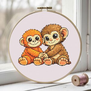 May include: A cross-stitch embroidery hoop featuring two cartoon monkeys. One monkey is orange, the other is brown, and they are holding hands. The embroidery is set against a light pink fabric, framed by a wooden hoop.