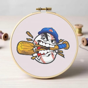 Może przedstawiać: Haft krzyżykowy w drewnianej tamborku przedstawia wściekłą piłkę do baseballa w niebieskiej czapce, gryzącą żółty kij. Piłka ma czerwone szwy na białym tle. Tamborek ma średnicę około 15 cm.