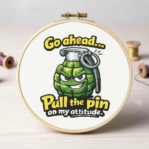 May include: Cross-stitch embroidery in a wooden hoop features a cartoon grenade with an angry face. The text reads "Go ahead... Pull the pin on my attitude." The design is in shades of green, gray, and yellow.