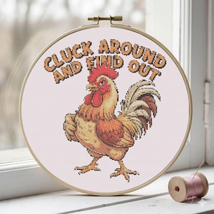 Può includere: Ricamo a punto croce con un gallo e la scritta "CLUCK AROUND AND FIND OUT". Il gallo è nei toni dell'arancione, del rosso e del giallo. Il telaio da ricamo è in legno e viene presentato su una superficie bianca. Diametro di circa 20 cm.