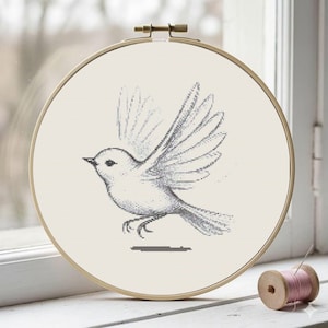 May include: A cross-stitch embroidery hoop featuring a detailed grayscale drawing of a bird in flight. The bird's wings are spread, and the design is set against a cream-colored fabric. A wooden spool of pink thread sits nearby.