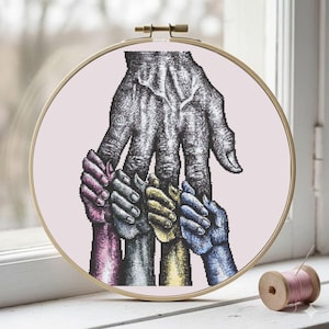 May include: Cross-stitch embroidery in a wooden hoop. The design features a large, detailed hand in grayscale, with smaller, colorful hands grasping it. The smaller hands are in shades of pink, gray, yellow, and blue. The background is a light pink.