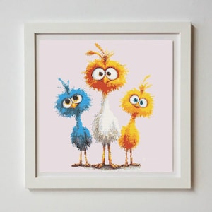 May include: A framed cross-stitch picture depicting three cartoon birds. The central bird is white, with a blue bird to the left and an orange bird to the right. They have large eyes and a fluffy texture. The background is a pale pink, and the frame is white.