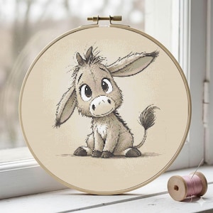 Pode incluir: Um bastidor de bordado em ponto cruz com um simpático burro de desenho animado com olhos grandes e orelhas caídas. O burro é em tons de cinza e bege, sobre um fundo bege. O bastidor de madeira é exibido numa janela branca.