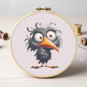 May include: A cross-stitch embroidery hoop featuring a cartoon bird with large, expressive eyes and an orange beak. The bird is primarily dark gray with lighter gray shading. The embroidery is set in a wooden hoop, with spools of thread in the background.