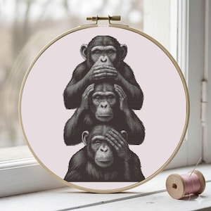 May include: A cross-stitch embroidery hoop showing three chimpanzees in the 'see no evil, hear no evil, speak no evil' pose. The monkeys are in black and white, set against a pale pink background. The hoop is round with a gold-coloured frame.