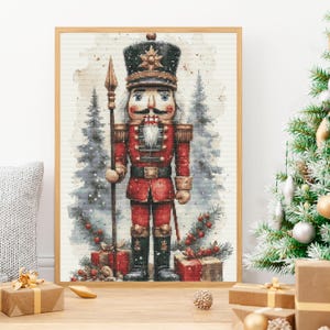 May include: A framed cross-stitch artwork depicting a nutcracker soldier in a red uniform with a tall black hat, holding a spear. The figure stands before snowy pine trees and wrapped gifts, rendered in a detailed, textured stitch pattern.
