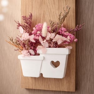 Könnte beinhalten: Wanddekoration mit einem weißen Pflanzgefäß in Herzform, gefüllt mit rosa und getrockneten Blumen. Das Pflanzgefäß ist auf einer rechteckigen Holzplatte befestigt und bildet eine charmante Blumendekoration.