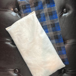 May include: Two rectangular fabric items. One is a cream-colored, soft pillow. The other is a plaid pattern in blue, black, and gray. The items are resting on a dark brown, tufted surface.