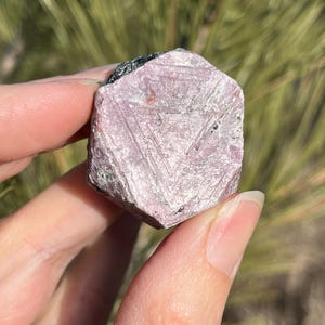 May include: A light pink, faceted gemstone with a geometric pattern and a rough texture. The stone is held in a hand. The background is blurred, with green and brown tones.