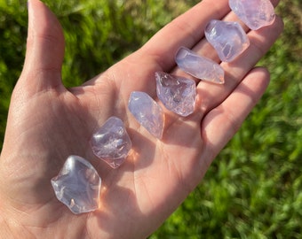 May include: A collection of six translucent, light purple, irregularly shaped stones. The stones are smooth and have a slightly iridescent sheen.