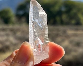May include: A clear quartz crystal point with a natural, rough texture. The crystal is held up against a blurred background of mountains, trees, and a blue sky. The crystal's facets catch the light, creating a sparkling effect.