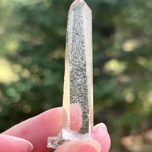 May include: A clear, elongated quartz crystal point with a hexagonal shape and a pointed tip. The crystal features a textured, frosted interior inclusion that runs vertically through the center, contrasting with the smooth, transparent outer facets.