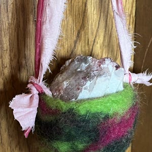 May include: A small, handmade pouch crafted from green, black, and magenta wool, holding a large, clear crystal. The pouch is suspended by pink fabric straps. The crystal has a rough, natural texture.