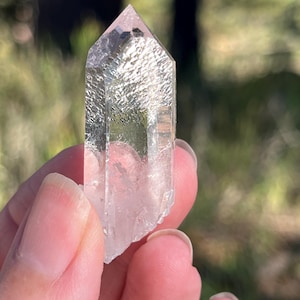 May include: A clear, six-sided quartz crystal point with a pointed termination. The crystal features a textured, frosted surface on its upper facets, transitioning into a transparent, smooth base with visible internal inclusions and fractures.