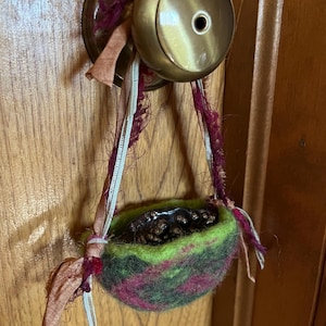 Peut inclure: Un petit panier en feutre vert et magenta, rempli d'objets brun foncé, est suspendu à un bouton de porte en laiton. Le panier est suspendu par des rubans roses, blancs et magenta. Le bouton de porte est monté sur une porte en bois.