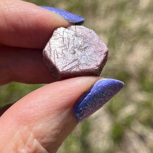 May include: A faceted, pinkish-red gemstone with a crystalline structure. The gem is held between two fingers with blue glitter nail polish. The stone has a hexagonal shape and appears to be a raw, uncut mineral.