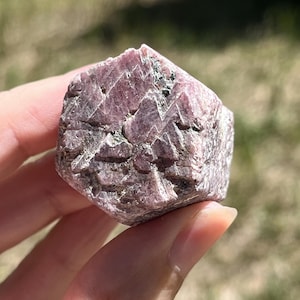 May include: A raw, hexagonal-shaped crystal specimen with a deep magenta and dusty rose coloration. The surface features a rough, textured appearance with visible crystalline facets, natural fissures, and small dark mineral inclusions throughout.