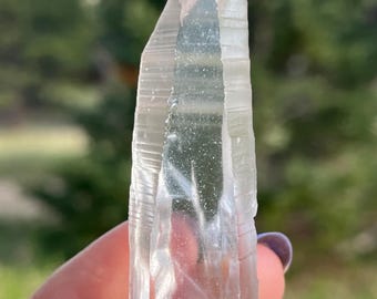 May include: A clear quartz crystal point, held against a blurred green background. The crystal is transparent with visible internal striations and a slightly frosted appearance. The crystal is approximately 4 inches long.