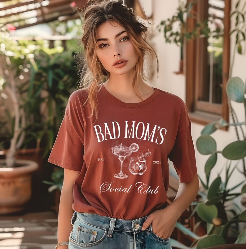 Peut inclure: Une femme portant un t-shirt rouille avec le texte "Bad Mom's Social Club" et un graphique blanc de deux cocktails. Le texte "Est. 2024" est &eacute;galement imprim&eacute; sur le t-shirt.