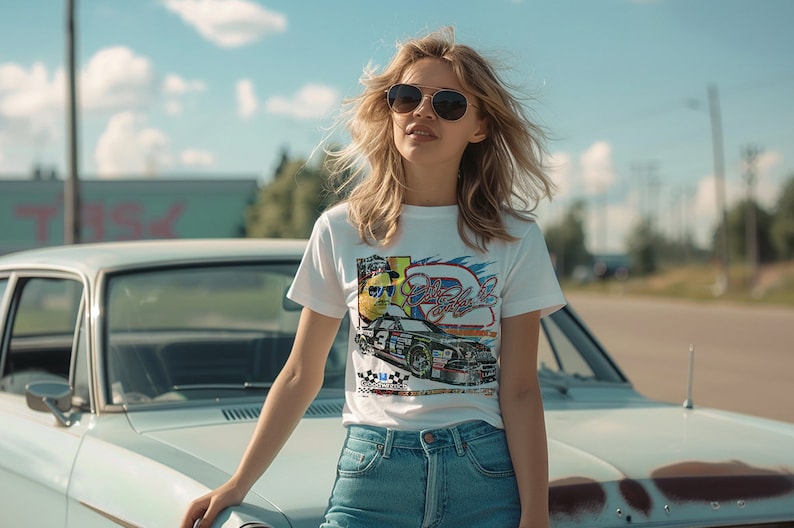 May include: A woman wearing sunglasses and a white t-shirt with a colorful graphic of a race car. The t-shirt has the text "Dale Earnhardt Jr." on it. She is standing next to a light blue vintage car.