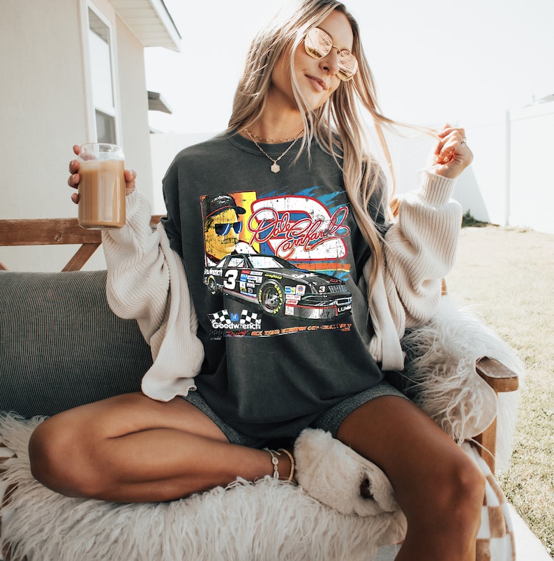 May include: A dark grey t-shirt with a vintage-style graphic of a race car driver and a race car. The graphic includes the text "Dale Earnhardt Jr." and "Goodwrench".