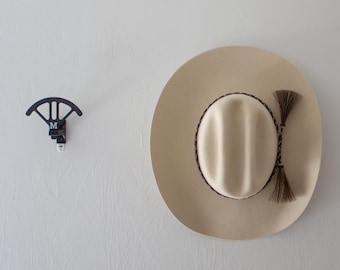 Könnte beinhalten: Ein weißer Cowboyhut mit braunem Band und einem geflochtenen Lederriemen, der vom Hutband hängt. Der Hut hängt an einer weißen Wand mit zwei schwarzen und weißen Haken.