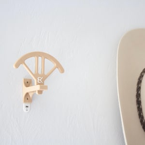 May include: A light-colored wooden hat rack mounted on a white wall. The rack has a curved top and a lower support, with a logo. A portion of a beige cowboy hat is visible to the right.
