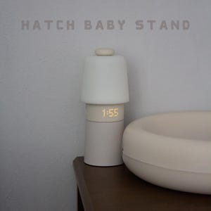 May include: A cylindrical Hatch Baby Stand lamp with a matte off-white finish, featuring a digital clock display showing 1:55 in yellow light. The lamp has a smooth, rounded top button and a wide, circular base, positioned next to a curved object.