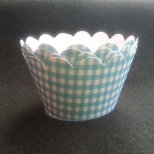 May include: A single cupcake liner with a scalloped edge. The liner features a blue and white gingham pattern on the outside and a white interior. The liner is designed to hold a cupcake.