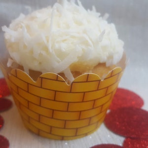 May include: A yellow cupcake wrapper with a brick pattern, topped with white coconut frosting. The cupcake is sitting on a white surface with red glitter confetti.