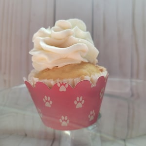 May include: A single cupcake with white frosting and a pink paw print liner. The cupcake sits on a clear glass cake stand. The frosting is swirled and fluffy, and the cupcake itself is golden brown.