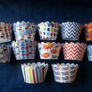 May include: A collection of superhero-themed cupcake liners in various patterns. Designs include cityscapes, zigzags, polka dots, and comic book exclamations like "ZAP!" and "POW!" Colors include red, blue, white, and yellow. The liners have a scalloped edge.