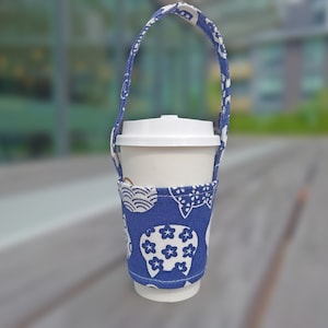 May include: A reusable coffee cup holder with a blue and white floral pattern. The holder has a matching handle and is designed to fit a standard coffee cup. The cup is white with a white lid.