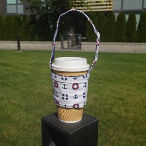 May include: A white fabric coffee cup holder with a blue and red anchor and life preserver pattern. The holder has a black strap and is holding a brown paper coffee cup.