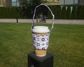 May include: A white fabric coffee cup holder with a blue and red anchor and life preserver pattern. The holder has a black strap and is holding a brown paper coffee cup.