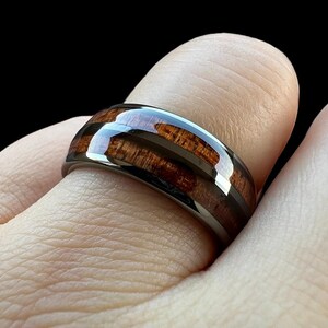 May include: A black tungsten carbide ring with a brown wood inlay. The ring has a polished finish and a beveled edge.