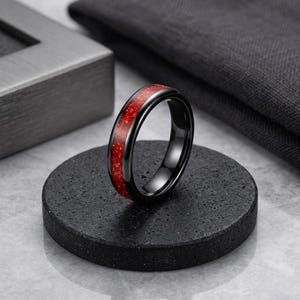 Op de afbeelding: Een zwarte ring met een levendige rode inleg. De ring staat op een donker, getextureerd oppervlak, dat de gepolijste afwerking benadrukt. De rode inleg heeft een gespikkelde uitstraling, wat visuele interesse toevoegt.