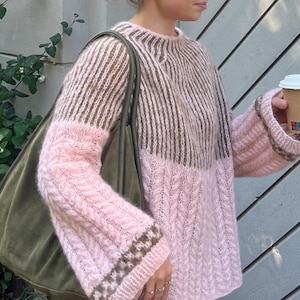 May include: A long-sleeve knit jumper featuring a colour-blocked design with a brown ribbed yoke and a pink cable-knit body and sleeves. The jumper includes a brown patterned band near the cuffs and is paired with a large olive green shoulder bag.