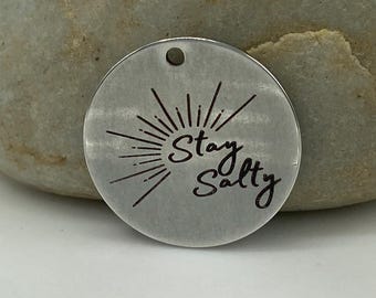 May include: A silver round charm with a sunburst design and the text "Stay Salty".