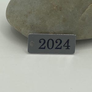 May include: A silver metal tag with a black number "2024" printed on it.