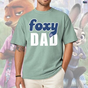 May include: A light green t-shirt with the words "foxy DAD" in a blue and white design. The shirt features a fox tail graphic. The background includes cartoon characters from the movie Zootopia.