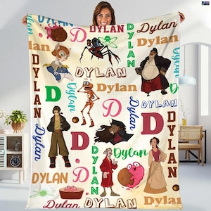 May include: A cream-colored blanket featuring cartoon characters and the name "Dylan" in various fonts and colors. The design includes the letter "D" and the word "Dylan" repeated throughout the blanket.