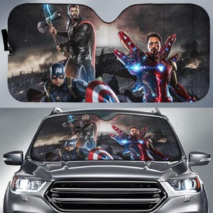 Op de afbeelding: Een zonnescherm voor de voorruit van een auto met een grafische print van Thor met een hamer, Captain America met een schild en Iron Man in een rood en goud pak. De achtergrond toont een donker, stormachtig slagveld met puin en wolken.