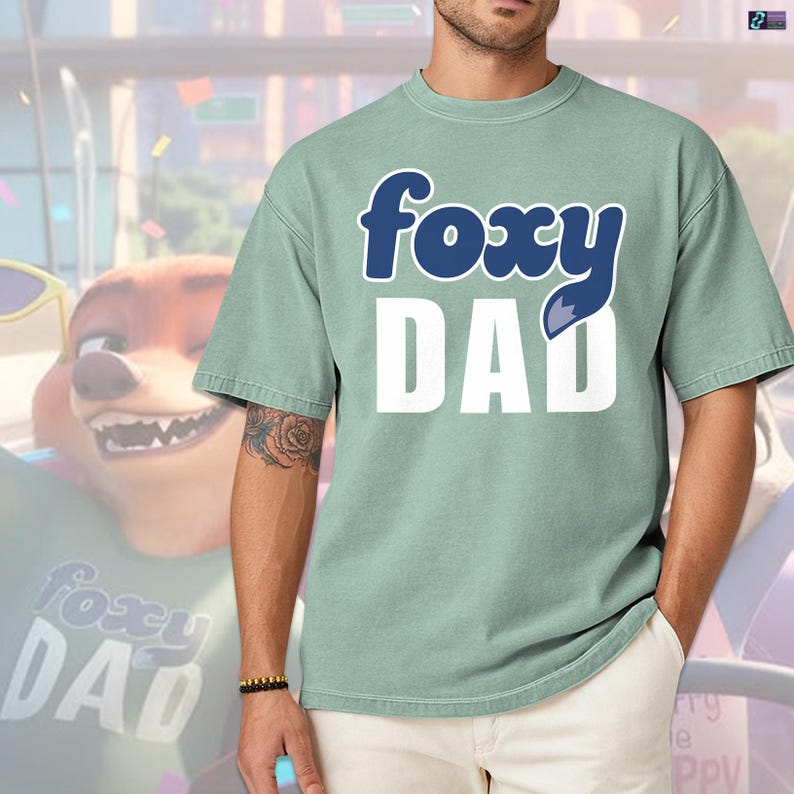 May include: A light green t-shirt with the text "foxy DAD" in white and blue. The word "foxy" is in a stylized font, and the letter "A" in "DAD" has a fox tail design. The shirt is a casual style.
