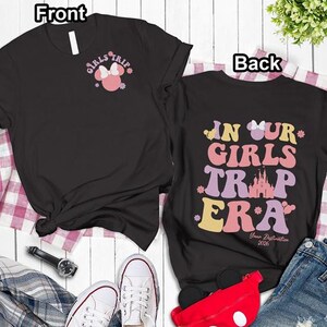 a pair of black t-shirts with a Disney-themed design, laid out on a white surface with a plaid cloth. The t-shirts have a front and back view, and the text "Girls Trip" and "In Our Girls" are visible on the front, along with the text "Your Reflection" and "Your Reflection 2020" on the back.