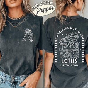 Könnte beinhalten: Dunkelgraues T-Shirt mit weißer Grafik eines Schlüssels und eines Hotelanhängers mit der Aufschrift "Lotus Hotel & Casino" auf der Vorderseite. Die Rückseite zeigt ein Lotusblumen-Design mit dem Text "You Will Never Grow Old Here" und "Lotus Las Vegas, Nevada."