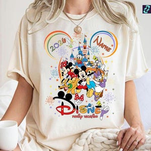 May include: Cream-colored t-shirt featuring a colorful Disney family vacation design. The graphic includes Mickey Mouse, Minnie Mouse, Donald Duck, Daisy Duck, and other characters. The design also includes the text "2020" and "Name" in a rainbow arch.
