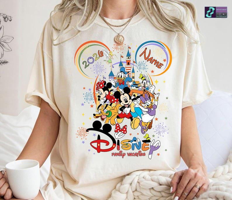 May include: Cream-colored t-shirt featuring a colorful Disney family vacation design. The graphic includes Mickey Mouse, Minnie Mouse, Donald Duck, Daisy Duck, and other characters. The design also includes the text "2020" and "Name" in a rainbow arch.